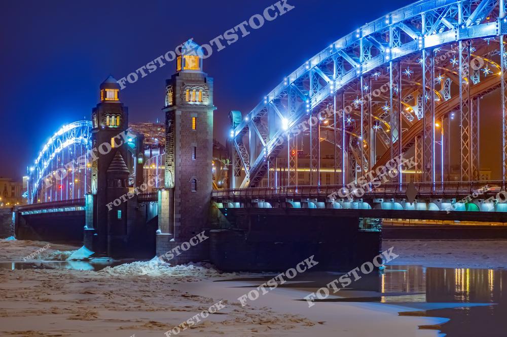 На фотографии изображен Большеохтинский мост в Санкт-Петербурге, освещенный яркой ночной подсветкой. Замерзшая Нева и льдины подчеркивают зимний характер пейзажа. Элементы архитектуры моста, включая массивные башни и арки, выглядят величественно и гармонично. Этот мост является не только транспортной артерией, но и одним из знаковых мест города.